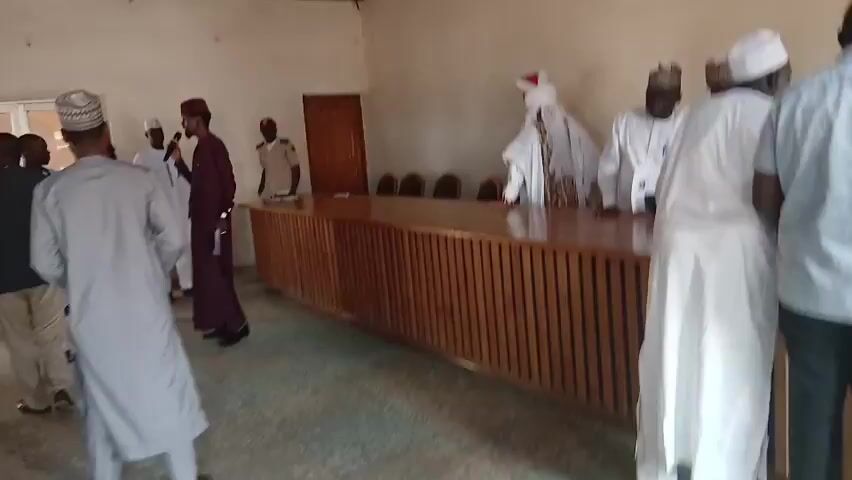 The video captures an indoor gathering of men in Sabon Gari North, Nigeria. The setting appears to be a meeting room or a similar communal space, indicated by a long wooden table with chairs behind it, a ceiling fan, and an air conditioning unit. The walls are painted a light color, and the ceiling has a distinctive textured pattern.

The subjects are predominantly men, many of whom are wearing traditional attire, including various styles of caps and robes, suggesting a cultural or religious context. One man in a dark purple robe and a red cap stands near the center, holding what appears to be a microphone, possibly addressing the group or participating in a discussion. Other men are gathered around, some in conversation, others observing. The lighting suggests it is daytime.

The activity seems to be a meeting, discussion, or gathering of some sort. The atmosphere appears to be formal but also engaged, with people interacting and participating. There is no visible text in the video.