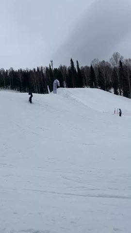 A lone snowboarder in dark clothing descends a snow-covered hill. At the top of the hill, there's a ramp with a flag bearing "EPARK".  A sparse forest flanks the hill, and a cloudy sky looms above. A mesh fence, marked with red, runs along the slope, and a couple of people are seen at the edge of the fence. The snowboarder briefly falls before continuing to ride down the hill.