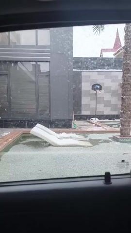 The camera is in a vehicle, looking through the window at a backyard with light snow falling. There is a small pool, partially iced over with pool furniture partially submerged and covered in snow. Beside the pool is a tall palm tree and a basketball hoop, with snow on the ground around the pole. There is a grey building with a large window and a grey brick wall next to it. There are wooden planks lying near the basketball hoop.