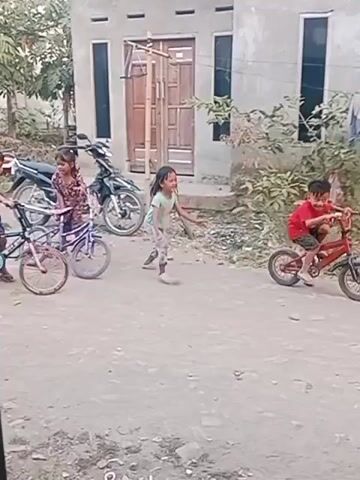 A group of children are playing outside near a house. One child is riding a bicycle. Another child is running. Two younger children are sitting on the ground. There is a motorcycle parked near the house. The ground is dirt and gravel.