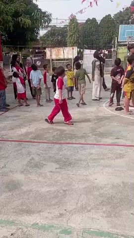 A group of children and adults are gathered on a paved outdoor court, possibly a basketball court, given the presence of a basketball hoop in the background. The ground is marked with red and white lines. Several children are wearing red and yellow shirts and red pants. Some adults are also present, including a woman wearing a black hijab. Flags and banners are strung across the top of the frame, suggesting a festive occasion. The children appear to be participating in some kind of game or activity, with some standing in a circle. One child in red pants is in the foreground, running. Another child, wearing a yellow shirt and red shorts, is standing in the center. The overall atmosphere seems to be a community event or celebration.