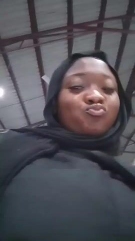 A woman wearing a black hijab and black jacket is looking at the camera and then looking up. She appears to be indoors in a large space with a metal ceiling.