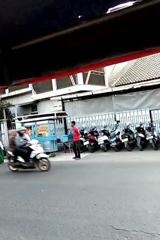 This is a short, slightly blurry video clip that appears to have been filmed from a moving vehicle, possibly a car or a pedicab, looking out at a street scene. The foreground is dominated by a dark, curved structure, likely the top edge of the vehicle the camera is in.

In the background, there is a street with several motorcycles parked in a row. To the left of the parked motorcycles is a food cart with a blue and white structure. Two people are standing near the food cart. One person, wearing a red shirt and dark pants, is facing away from the camera, possibly interacting with the person at the cart. Another person, wearing a patterned dress and a hijab, is facing the person in the red shirt and appears to be conversing. Further left, a person in a white outfit and a pink headscarf is visible, appearing to be on a bicycle.

Throughout the clip, several vehicles are seen moving. A white scooter with two riders passes by in the middle of the street. Later, a black SUV drives past, obscuring the view of the motorcycles and the building behind them. Another motorcycle, with a rider wearing a light-colored jacket and a dark helmet, is also seen moving. The building behind the parked motorcycles has a white facade with multiple windows. The overall impression is of a busy street, likely in an urban or suburban area, with activity around a food stall and traffic on the road.