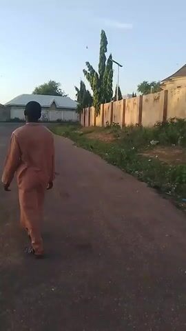 The video shows a view from behind two individuals walking away from the camera down a paved road. The first individual, closer to the camera on the left, is wearing a dark shirt and trousers and carrying a white cord in their right hand. The second individual, to the right, is wearing a light brown robe and appears to be carrying a dark object in their right hand. The road is bordered on the right by a low, light-colored wall topped with concrete posts, and beyond the wall are some buildings with pitched roofs and tall, thin trees. On the left side of the road, there are also buildings visible behind a fence. The sky is clear and blue, suggesting it is daytime, likely morning or late afternoon due to the soft lighting. The overall environment appears to be a residential area.