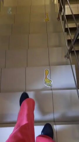 The video shows a set of stairs with decorative footprints placed on them. A person wearing red pants and black shoes is walking down the stairs. The footprints are yellow with an orange outline and appear to be positioned to guide someone down the stairs. The stairs have a tiled surface and a metal railing. The lighting suggests an indoor environment.