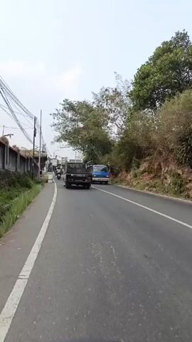 A POV shot from a moving vehicle on a two-lane road. On the left side of the road, there is a sidewalk, a low wall, buildings, and utility poles. On the right side of the road, there is a steep, vegetated embankment. Several vehicles are visible on the road. In the foreground, the road is asphalt with a white line on the left edge. In the midground, a black pickup truck is traveling away from the viewer, followed by a blue van. Further down the road, a white box truck is visible, followed by a line of motorcycles and a black car. In the background, more vehicles and buildings are visible, along with trees and the sky. The weather appears to be overcast.