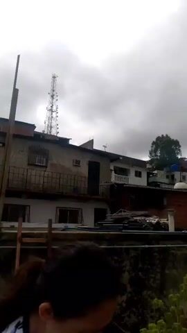 This video shows a city scene with buildings and a cloudy sky. In the foreground, there are some clothes hanging on a clothesline. In the background, there are several buildings, some of which appear to be unfinished. There is also a tall communication tower visible. In the distance, more buildings are scattered across hills. A person is seen in the lower left corner of the frame, wearing a t-shirt and pants, and they appear to be stretching or exercising. The sky is overcast with gray clouds.
