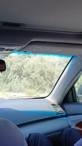 The video shows the interior of a car driving down a road. The view is from the backseat, looking forward. Trees and foliage can be seen through the windshield. The dashboard of the car is visible, including an air vent and part of the steering wheel. There are two people in the front seats, their legs and torsos visible from the shoulders down. The person in the passenger seat has on dark blue pants. The person in the driver's seat has on dark blue pants and a dark blue shirt. The car appears to be moving at a moderate speed, as the scenery outside is blurred. The sun is shining, casting light on the interior of the car.