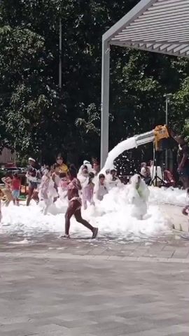 This is a video of a foam party. Several children are playing in a large pile of white foam generated by a foam machine. The foam machine is yellow and mounted on a stand. A person is operating the machine. Several adults are visible in the background, some watching the children, and some standing near the machine. The party appears to be taking place outdoors in a paved area with trees in the background and a covered structure overhead. The children are wearing swimsuits and appear to be having fun.