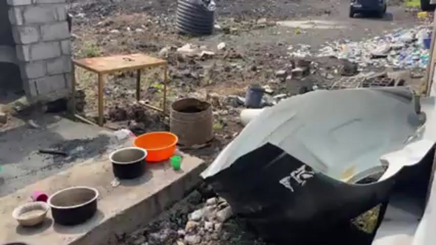 The scene shows a cluttered outdoor area with a table, various containers, and a large, damaged piece of what appears to be a vehicle part. The ground is covered in dirt, debris, and scattered trash. In the background, there's a water tank and a vehicle parked in the distance. The overall environment seems unkempt and possibly located in a less developed area.