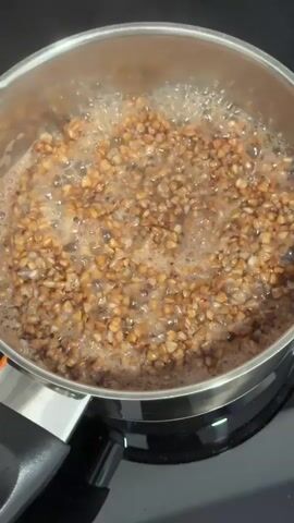 Buckwheat is boiling in a pot on a stovetop. The pot is made of silver metal and has a black handle. The buckwheat is a light brown color and is bubbling vigorously. The stovetop is black.