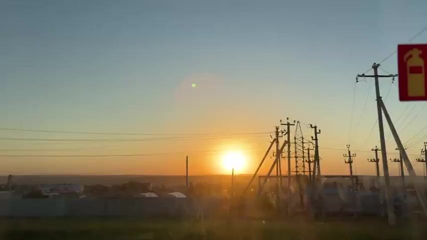 The video shows a sunset over a landscape with power lines and buildings. The sun is a bright orange ball on the horizon. The sky is a gradient of blue to orange. There is a red sign partially visible on the right side of the frame. The view is from behind a dirty window. A voice can be heard speaking in the background.
