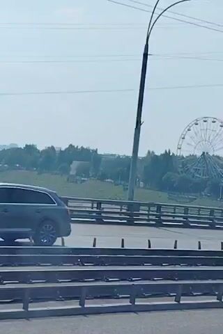 A white box truck is driving on a road. In the background, there is a Ferris wheel and trees. The sky is clear and blue.