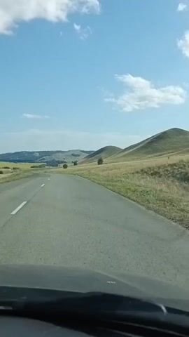 A first-person perspective video shows a car driving on a paved road through a rural landscape. The road is asphalt with white dashed lines on the left side. The landscape is characterized by rolling hills covered in dry, yellow grass. There are scattered trees on the hillsides and in the distance. The sky is blue with scattered white clouds. The video appears to be taken on a sunny day. The car's dashboard is visible at the bottom of the frame. There are no people visible in the video.