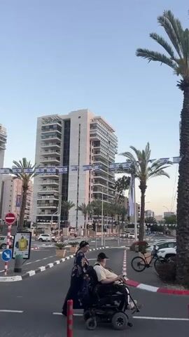 The video captures an urban street scene in Kiryat Motzkin, Israel, during the late afternoon or early evening, indicated by the clear sky transitioning to soft pink and orange hues at the horizon.

In the initial frames, an older man seated in a motorized wheelchair is being pushed by a woman across a crosswalk in a multi-lane street. The woman wears a dark patterned dress and head covering. The street is lined with numerous parked cars, several tall palm trees, and modern, multi-story buildings.

As the camera pans to the right, more parked cars are visible, alongside additional palm trees and buildings. Prominently strung across the street, between buildings and lampposts, are rows of Israeli flags, featuring the blue Star of David. A large billboard with an advertisement is visible on the left side of the frame. A street light is illuminated, further suggesting the approach of dusk. The scene depicts a typical city environment with pedestrian activity and parked vehicles under a fading sky.