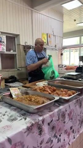 A bald man wearing a blue t-shirt and a black apron is standing behind a counter in what appears to be a butcher shop or food market. He is holding a bright green plastic bag in his right hand and is in the process of handing it to someone off-camera, whose hand is visible holding the other side of the bag. In front of the man, on the counter, are several stainless steel trays filled with raw meat, some of which appears to be marinated. The counter is covered with a floral-patterned tablecloth. In the background, there is a white wall, a scale, a refrigerator, and some religious icons. The man is looking towards the person he is interacting with.