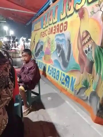 A child is sitting on a small green plastic chair at an outdoor food stall. The child is wearing a maroon jacket, yellow shorts, and sandals. The food stall has a large sign with colorful drawings of a rooster, fish, and other items. The sign is in Indonesian and advertises "Ayam Geprek, Lele Goreng Crispy, Nasi Bebek."  Other people are visible in the background, walking around. The setting appears to be a nighttime street market.