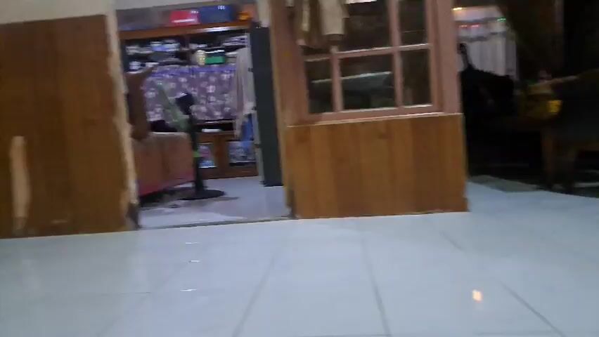 The video presents two distinct interior scenes.

The first scene (0:00-0:01) is a wide shot from a low angle, showcasing a domestic setting. The camera is aimed towards a doorway, revealing another room with shelves filled with various items, and what appears to be an exercise machine or treadmill. The walls in the foreground have wood paneling, contrasting with a light-colored floor. A potted plant is visible on the left side of the frame, and a window with a wooden frame is partially seen on the right.

The second scene (0:01-0:05) features a young woman in a close-up, selfie-style shot. She has long dark hair, light-colored contact lenses, and is wearing a black top. She is positioned against a background of soft furnishings, including pink, blue, and red blankets or pillows. A white and blue spray can, with the word "ROOM" visible on its label, and a pink strap are also seen in the background.

The woman performs several actions:
1. Initially, she looks directly at the camera with a neutral expression.
2. She then opens her mouth and sticks out her tongue, which is notably bifurcated (split down the middle).
3. Following this, she makes a peace sign with her right hand, winks, and smiles playfully, engaging with the camera.

The overall mood is light and playful. The setting is likely a bedroom or personal space within a home in Mempawah, Indonesia.