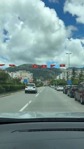 A view of a road from within a moving car, looking down a straight, multi-lane road in what appears to be a European city, with mountains in the background. Red flags with a gold emblem are hung across the road at regular intervals. Several cars are on the road, traveling in both directions. On the right side of the road, cars are parked along the curb. A billboard stands on the left side of the road, displaying a photo of a man in a suit. Traffic lights can be seen ahead, showing green. The sky is cloudy with patches of blue visible. The camera is static within the car, providing a forward-facing perspective of the drive.