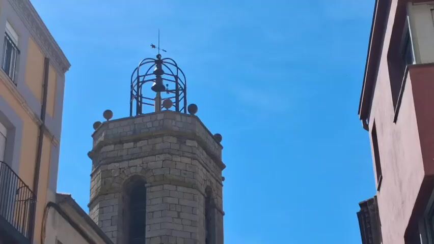 The video shows a street view in a town, with a bell tower and clock as prominent features.  The camera pans down from the top of the tower, showing the clock face which reads almost 11:50,  then moving to show the church's brick façade and arched entrance. Several people are walking in the street, and there are buildings on either side, painted in various colors including peach and pink. A stop sign is visible on a pink building.  The sky is clear and blue.  A bird flies overhead in several shots.