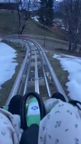 A first-person perspective video shows someone riding a single-rail alpine coaster. The camera is positioned low, showing the rider's legs and the coaster car. The tracks are metal and silver-colored, winding down a slight hill. There are patches of snow along the sides of the tracks, suggesting a winter or early spring environment. Bare trees and a few evergreen trees are visible in the background. The sky is overcast and the lighting is muted. The person riding the coaster is wearing dark shoes and light-colored pants with a floral pattern. The coaster car appears to be green and black.