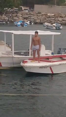 A young boy, shirtless and wearing dark swim trunks, stands on the edge of a white boat with a red stripe. He leaps into the water, creating a splash. The boat, named "MI LUCERITO" with "LA GOLFA" also visible on its side, is docked near other boats in a harbor. In the background, a rocky shoreline is visible with a town and hills beyond. The water is calm and appears to be in a tropical or coastal setting. The lighting suggests it is daytime.