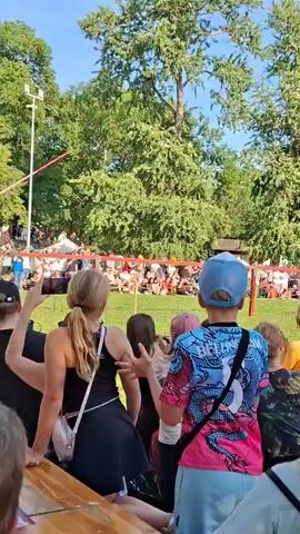 A jousting tournament is taking place outdoors. Two knights on horseback are charging at each other, with lances extended. A crowd of spectators is watching from behind a barrier, some of whom are clapping and cheering. The event is set against a backdrop of lush green trees and a clear blue sky.