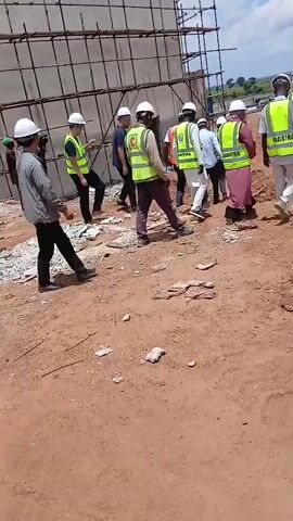 A group of people in construction safety gear are walking on a dirt construction site. They are wearing hard hats and high-visibility vests. One person is wearing a pink garment under their vest. In the background, there is a large building under construction with scaffolding and a crane. The sky is partly cloudy.