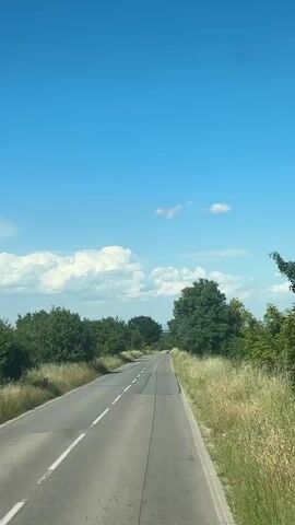 The video shows a view from a vehicle traveling on a paved road through a rural landscape on a clear day with a bright blue sky and scattered white clouds. The road has two lanes with a broken white line in the center and solid white lines on the edges. Tall, dry grass lines the sides of the road, interspersed with lush green trees and bushes. The road curves gently to the right as it goes uphill. There are no people or other vehicles visible in the frame. The overall scene is serene and natural.