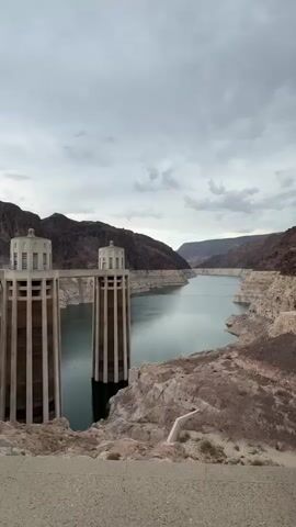 The video shows a view of a large body of water behind a dam. Two prominent concrete towers with circular structures on top stand in the foreground on the left side of the frame. The water is calm and reflects the cloudy sky. The surrounding landscape consists of steep, rocky hills and mountains, with visible evidence of water level marks on the dry banks, indicating a lower water level. The dam itself is partially visible in the distance across the water. The overall impression is one of a vast, natural landscape dominated by the man-made structure of the dam and the surrounding geology.