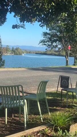 The video shows an outdoor seating area with two light green plastic chairs in the foreground. To the right, there is a small black sign with white text that reads "NORTH BEACH CAFE". Beyond the seating area is a paved road, and beyond that is a wide, calm body of water, likely a river or bay. Several boats, including a sailboat and a white motorboat, are visible on the water. The background features lush green trees and hills under a clear blue sky. The scene appears peaceful and scenic, suggesting a waterfront cafe setting.