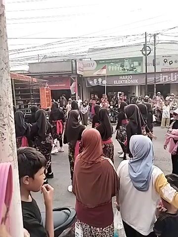 A group of people, mostly women wearing hijabs and traditional outfits, are dancing in the street. They are surrounded by a crowd of onlookers, some of whom are holding cameras or phones. The street is lined with buildings that appear to be shops. The sky is overcast. There are motorcycles parked along the side of the street. The overall atmosphere seems festive.