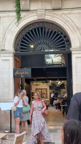 The video begins with a close-up view of an arched doorway, adorned with an intricate metal grate. Below the arch, a sign with the word "komera" in elegant script is visible. Two women stand near the entrance, one in a white t-shirt and denim shorts, the other in a patterned dress. A man with his back to the camera stands in the foreground, looking towards the entrance. The camera then pans up to reveal the upper floors of the building, showing a balcony with a wrought-iron railing and a window with closed shutters. The architecture appears to be old, with weathered stone walls. As the video progresses, the camera zooms out slightly, revealing more of the street scene. Several other people are visible in the background, including some seated at tables outside the establishment. The overall impression is of a bustling European street in front of a restaurant or cafe.