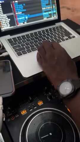 Here's a description of the video:

The video shows a DJ using a laptop and DJ controller at an outdoor event. The laptop screen displays DJ software. A smartphone is connected to the DJ controller. The DJ is wearing a watch on their left wrist and is using the laptop's trackpad with their right hand. In the background, people are walking around. A woman with red hair, wearing a white skirt and black boots, stands near the DJ setup. The ground is red dirt. The setting appears to be a casual outdoor party.