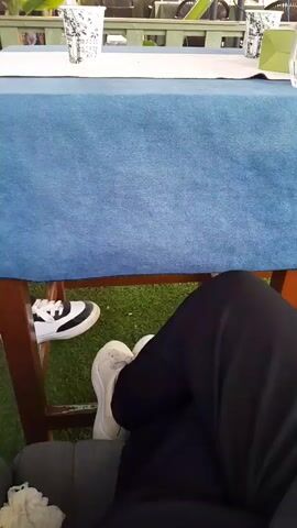 The video shows a person's legs and feet wearing black pants and white sneakers. The person is sitting under a blue tablecloth that is on a wooden table. To the left of the person's feet, another pair of black and white sneakers is visible. The background shows an outdoor seating area with tables and chairs, as well as some greenery and plants. The lighting suggests it is daytime.