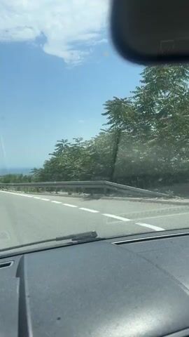 The video shows a car driving on a mountain road on a sunny day. The road has a metal guardrail on the side, and there are trees and mountains visible in the distance. The sky is blue with some clouds. The music is loud and can be heard.