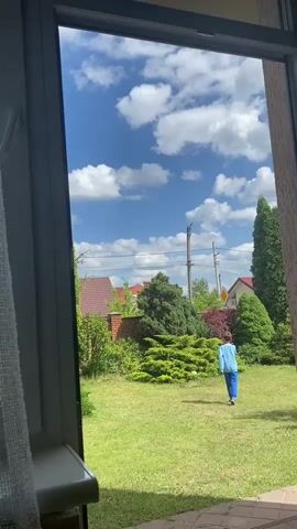A young boy wearing a blue tracksuit is playing in a backyard with green grass. He is throwing a white ball in the air and catching it. The backyard is surrounded by trees and bushes, and there are houses in the background. The sky is blue with white clouds. The video is taken from inside a house, looking out through a window.