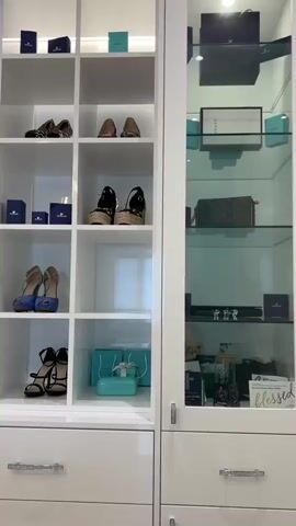 The video shows a closet with shelves displaying shoes and boxes. The shelves are white and illuminated by lights from above. On the right side of the closet is a glass-enclosed cabinet with more shelves. The cabinet contains handbags, boxes, and other accessories. The video pans across the shelves, showing different pairs of shoes and various boxed items. The shoes appear to be high heels and sandals, in different colors and styles. Some of the boxes are teal, suggesting they may be from a luxury brand like Tiffany & Co. The overall impression is of a well-organized and luxurious closet.