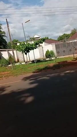 The video shows a street view of what appears to be a residential area.  The camera is moving, giving the impression of driving or riding along the road.  In the beginning, there is a large house with a gate and a tall wall around the property.  As the video progresses, the buildings transition to smaller, more modest structures.  There's a glimpse of what looks like a small shop or stand where clothes are displayed. The sky is visible, with some clouds.  The road has a white line marking the lanes. There are some trees along the side of the road.