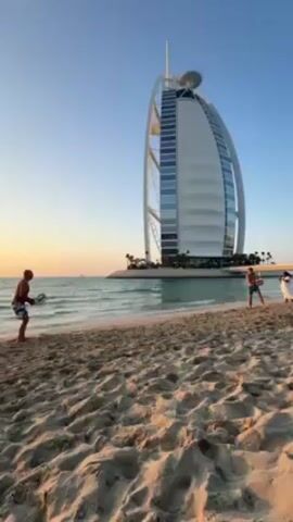 The video shows two people playing beach tennis on a sandy beach with the Burj Al Arab hotel in the background. The sky is clear and the sun is setting, casting a warm glow on the scene. The waves are gentle, and there are a few other people visible in the distance. The two individuals are holding rackets. They are hitting a ball.