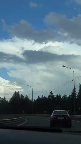 A view from inside a vehicle, looking out through the windshield at a highway.  A dark sedan is visible ahead on the road.  Streetlights and a line of trees line the sides of the highway.  The sky is blue with scattered clouds.  The windshield wipers are partially visible in the foreground.