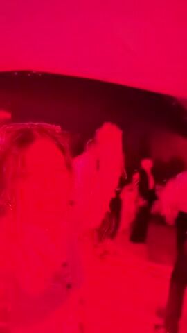 A woman with dark, curly hair is seen in the foreground of the video, illuminated by a bright pinkish-red light. She is wearing a white top and appears to be singing or shouting. Her mouth is open, and her eyes are wide. Her right arm is raised in the air. In the background, other people are visible, also in motion and under the same colored lighting. Some are blurry figures dancing or interacting. The environment suggests a party or a nightclub setting. The video is shaky and appears to be shot from a mobile device.