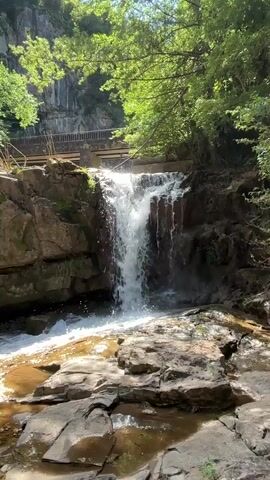 A waterfall cascades over rocks into a shallow pool. Trees and cliffs surround the waterfall, and a bridge can be seen in the background. The water is clear and flows rapidly. The sun shines through the trees, creating dappled light on the rocks and water. The scene is peaceful and natural.
