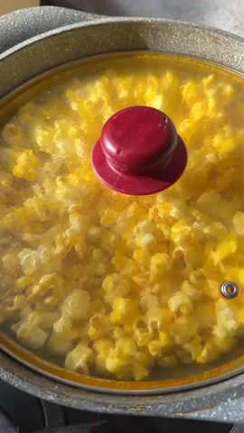 The video shows popcorn popping in a pan with a glass lid. The lid has a red knob in the center. The popcorn is yellow and fluffy. The pan is gray and has a textured outer surface. The lighting suggests the popcorn is being made in natural light, possibly near a window. The audio consists of the popping sounds of the popcorn.