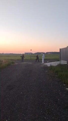 This is a vertical video of three people playing volleyball in a field at sunset. The sky is a pale orange and pink. The people are silhouetted against the sky. The person on the left is wearing a dark shirt and pants. The person in the middle is wearing a dark shirt and pants, and is jumping to hit the ball. The person on the right is wearing a white outfit. The ground is covered in gravel. There are some buildings in the background. The video is 5 seconds long.