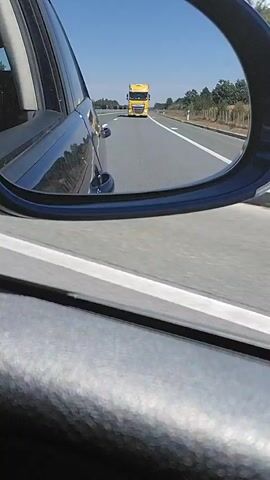 The video shows the view from inside a car on a highway. The camera is positioned to capture a side mirror and the car's interior. The side mirror reflects a bright yellow truck driving on the highway in the distance. The highway appears to be a multi-lane road with clear blue skies and trees lining the sides. The car is in motion, as evidenced by the blur of the road in the foreground and the movement of the truck in the mirror. The overall scene suggests a typical daytime drive on a highway.