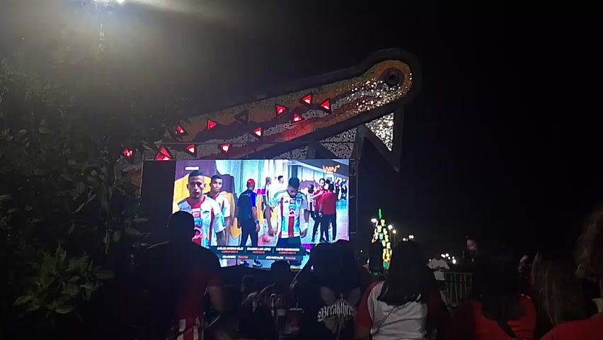 The video shows a crowd of people at an outdoor event at night. In front of the crowd is a large screen displaying a video of several men. Above the screen is a decorative archway with red lights. Many people in the crowd are holding up their phones, presumably to record or take pictures. The crowd seems to be focused on the screen, suggesting they are watching a performance or event.