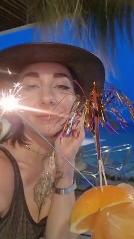 A woman wearing a hat and a dark tank top holds a sparkler in her right hand. She has tattoos on her left arm and chest. She is looking at the camera and smiling. To her right is a cocktail with an orange slice and a decorative umbrella. In the background, the sky is dark blue, suggesting it is evening. There is a blurry figure of a man sitting at a table in the background. Palm fronds are visible at the top of the frame.