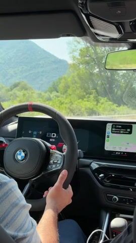 A close-up shot from inside a car shows a person driving. Their hands are on a black Alcantara-wrapped steering wheel with a red stripe at the top and a BMW logo in the center. A digital dashboard displays "80" prominently. To the right, an infotainment screen shows a map application via Apple CarPlay. Through the windshield, a tree-lined road is visible with green mountains in the distance. Another car is ahead on the road. The weather appears sunny.