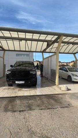 The video shows an outdoor self-service car wash on a bright, sunny day with a clear blue sky and scattered white clouds.

In the foreground, a large, black RAM pickup truck is parked in a car wash bay, being cleaned. The truck has a distinct "RAM" emblem on its grille and a Russian license plate, "с570сс 99," indicating the Moscow region.

A man, wearing a green plaid shirt, purple pants, and a dark cap, is standing to the right of the truck. He is actively washing the front left side of the vehicle with a high-pressure spray wand, directing water onto the truck's fender and wheel.

The car wash facility features bays with tiled floors, drainage grates, white partition walls, and overhead metal-framed canopies with translucent panels, providing shade. To the right of the black truck, a white sedan is partially visible in an adjacent car wash bay.

In the background, various buildings can be seen, including a modern-looking white commercial or industrial building, and further back, residential structures. A streetlight is also visible in the distance. The ground outside the car wash bays is paved with asphalt or concrete. The overall scene suggests a routine car maintenance activity taking place in Мокшино, Russia.